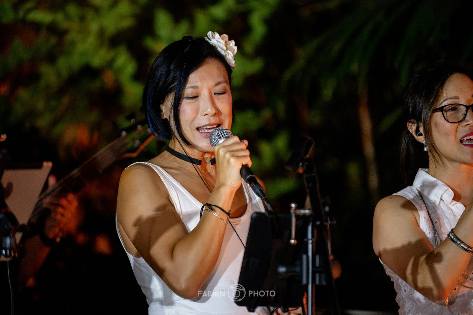 A woman with black hair, wearing a white sleeveless top and floral headband, singing into a microphone during a performance at night, with microphone stand and greenery in the background.