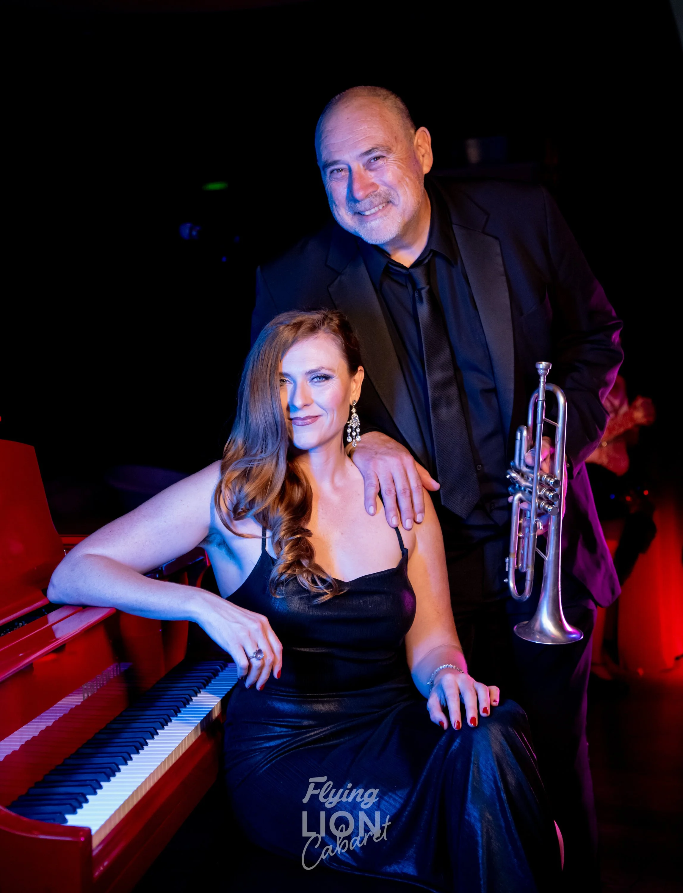 A man in a black suit with a trumpet standing behind a seated woman in a black dress, with a red piano beside her, in a dark room with colorful lighting. Weddings. Corporate Events. Private Parties. City Festivals. Tribute Shows. Live Band Burlesque.