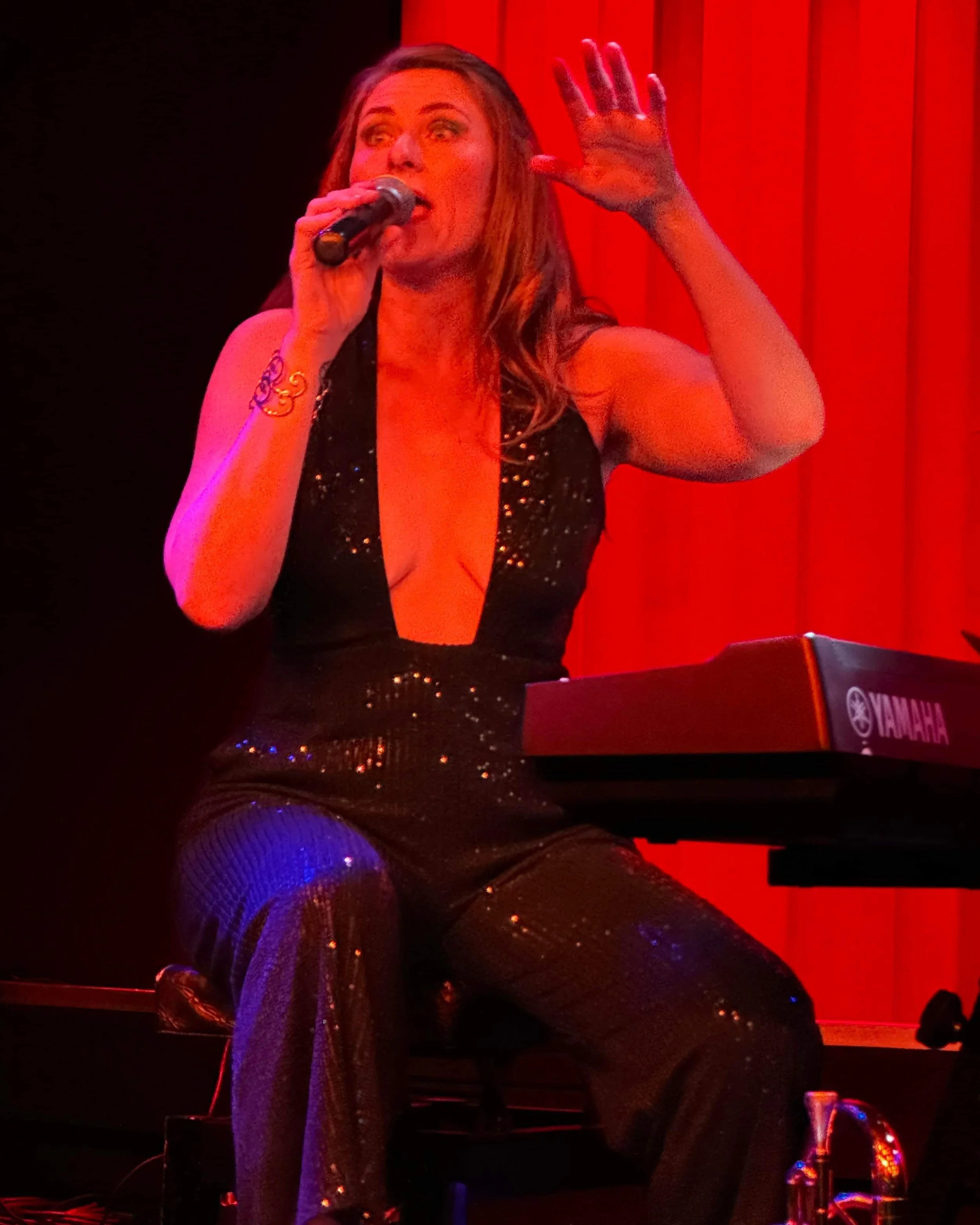 A woman singing into a microphone while playing a Yamaha keyboard on a stage with red curtains in the background. Weddings. Corporate Events. Private Parties. City Festivals. Tribute Shows. Live Band Burlesque. Tributonic.