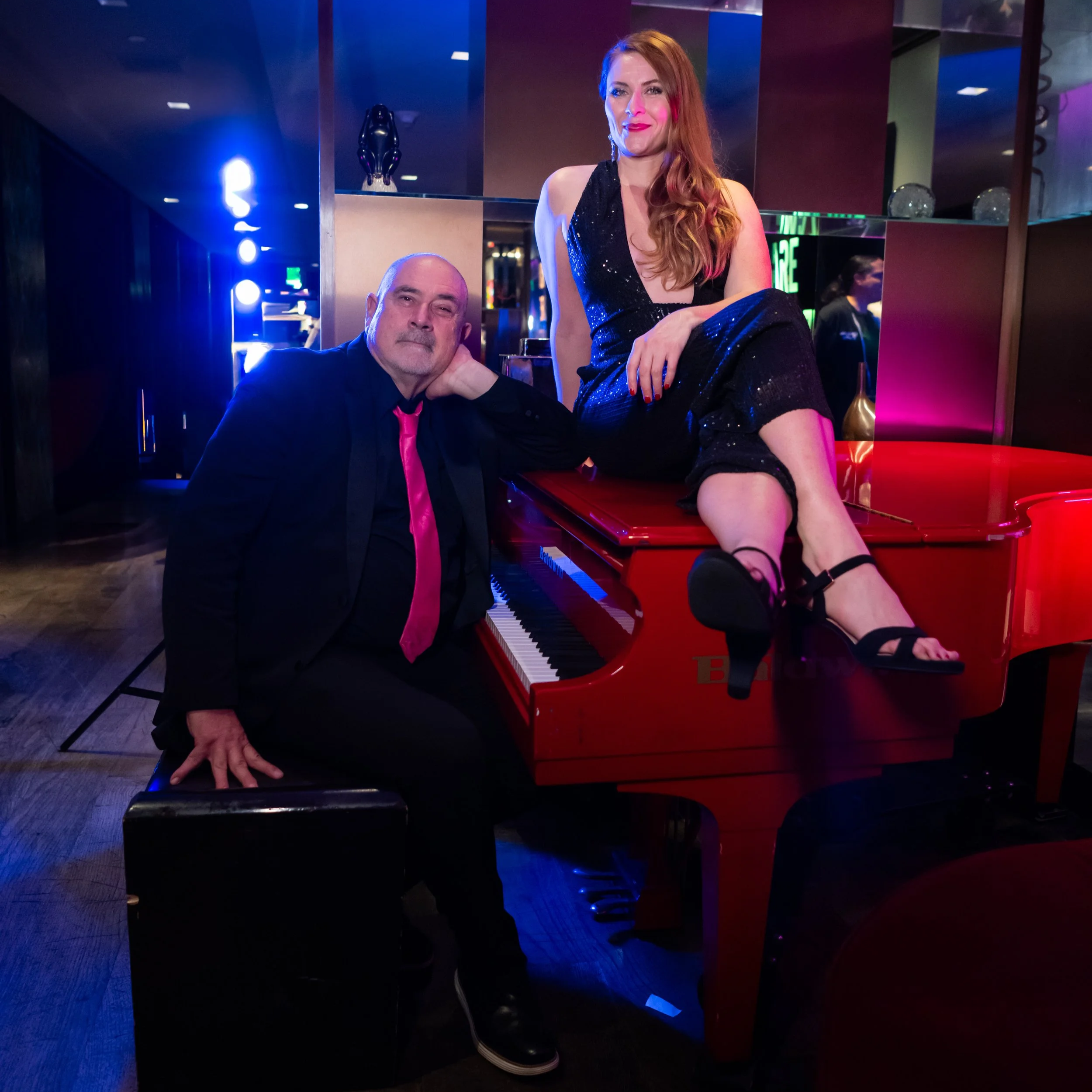 A man and a woman at a social event, the woman is sitting on a red piano wearing a black dress with sequin details, and the man is sitting beside her in a black suit with a pink tie. The woman has long red hair and is wearing black high heels, the man is bald with a goatee. The background features dim lighting with colorful lights and a modern, elegant interior.