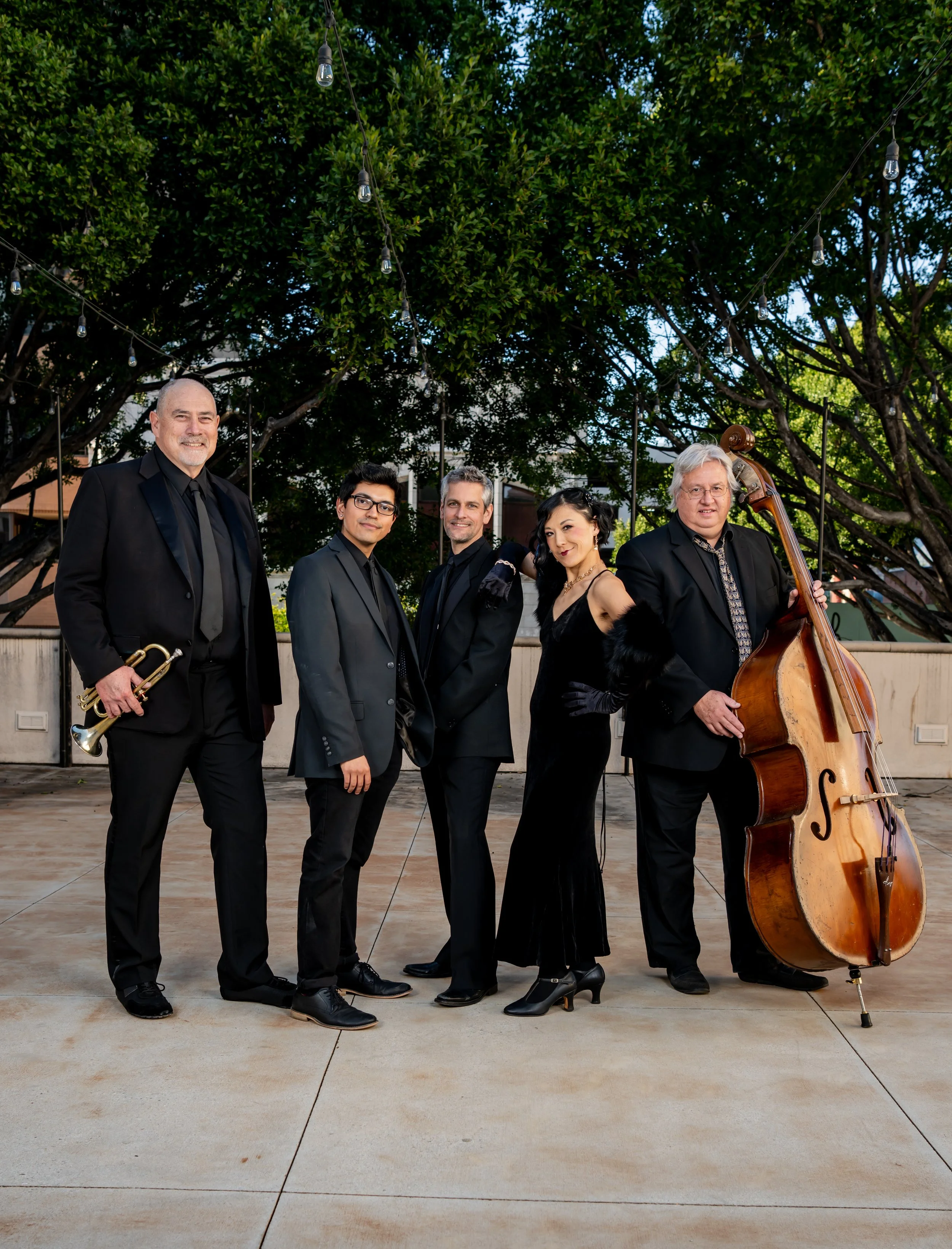 A jazz band performing outdoors, consisting of five members in black suits, with two holding musical instruments: a trumpet and a double bass. The background includes green trees, and string lights are hanging overhead.