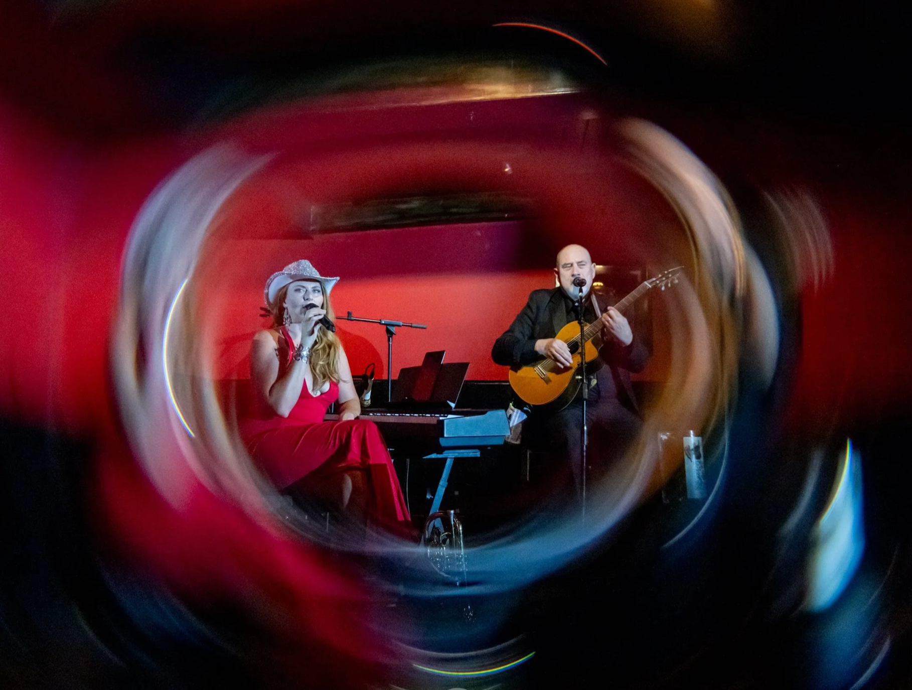 Two musicians performing on stage, a woman in a red dress and cowboy hat singing into a microphone and a man with a shaved head playing an acoustic guitar, with a red background.