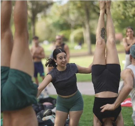 A woman assisting a person with an upside-down yoga pose in a park with other people practicing yoga nearby.