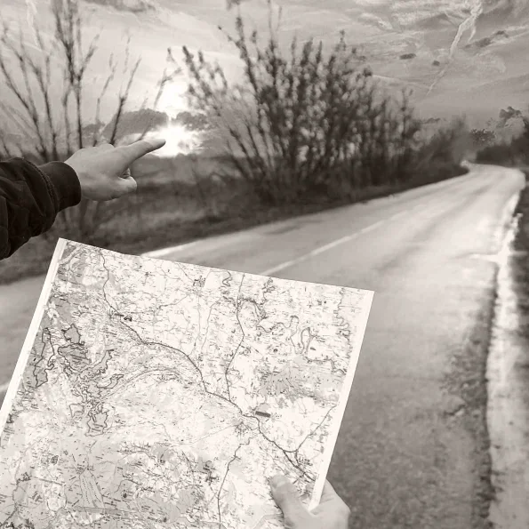 A person holding a map outdoors during sunset or sunrise, with one hand pointing towards the sky and the other holding the map near the road.