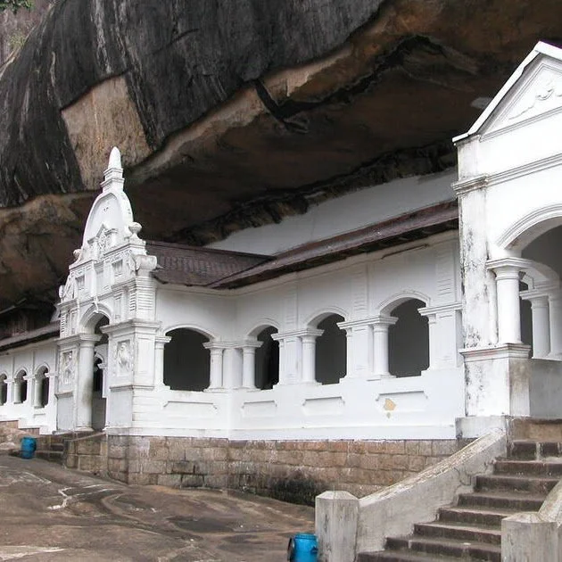 White building with arches and a steeple, built underneath a large rock overhang.