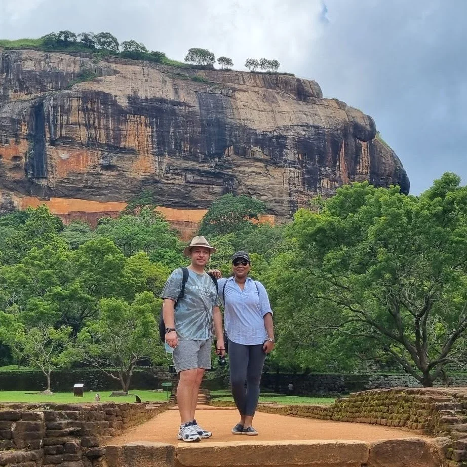 A man and a woman standing on a stone pathway in front of lush green trees and a large rocky cliff, with a partially cloudy sky above.