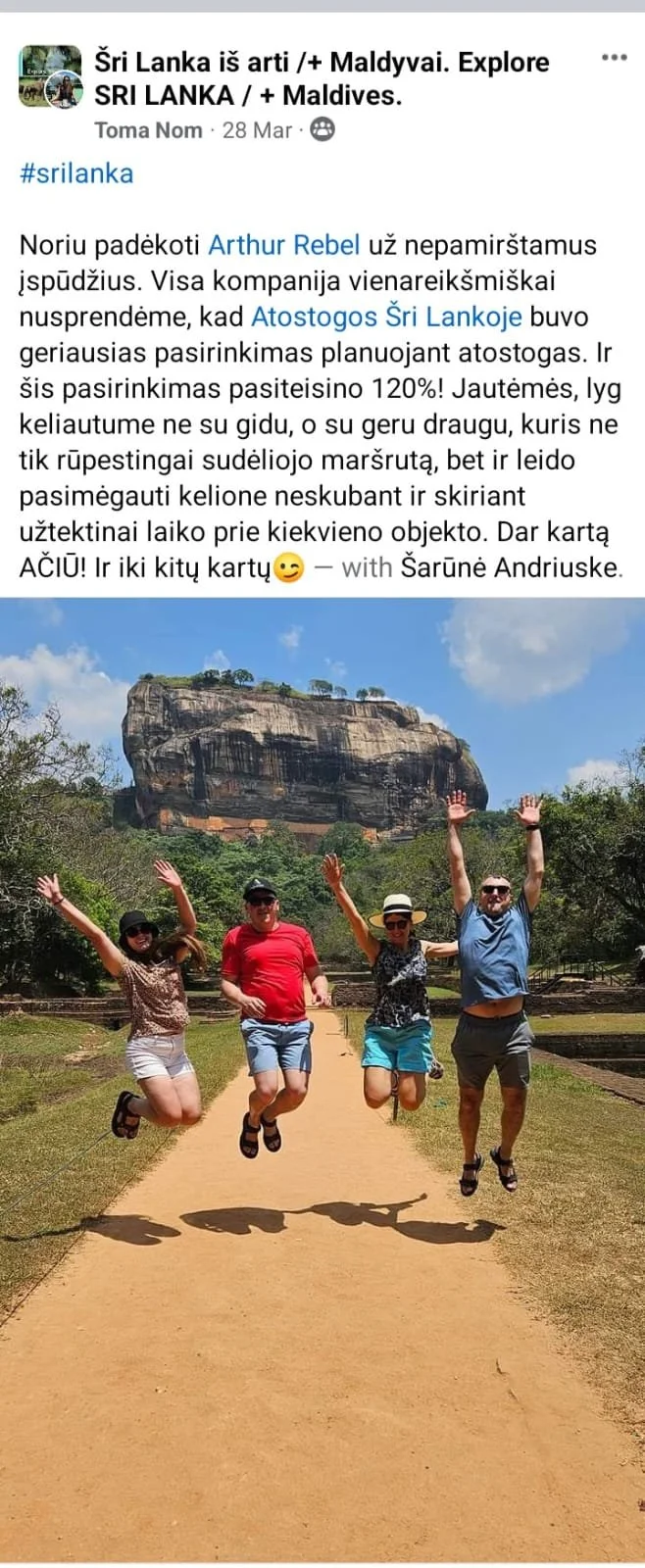 Four tourists jumping on a dirt path with a large rock formation in the background. They are wearing summer clothing and sunglasses, and the sky is partly cloudy.