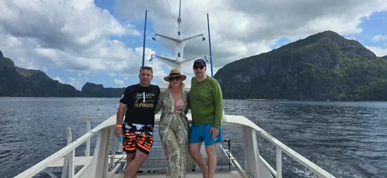 Three people standing on a boat in a scenic water setting with mountains in the background, dressed in casual summer clothing.