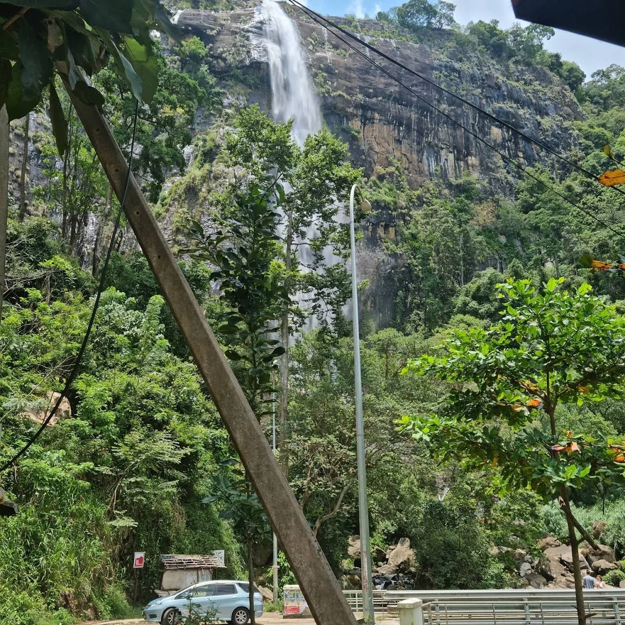Tall waterfall cascading down a rocky cliffside surrounded by dense greenery, with a road and parked vehicles in the foreground.