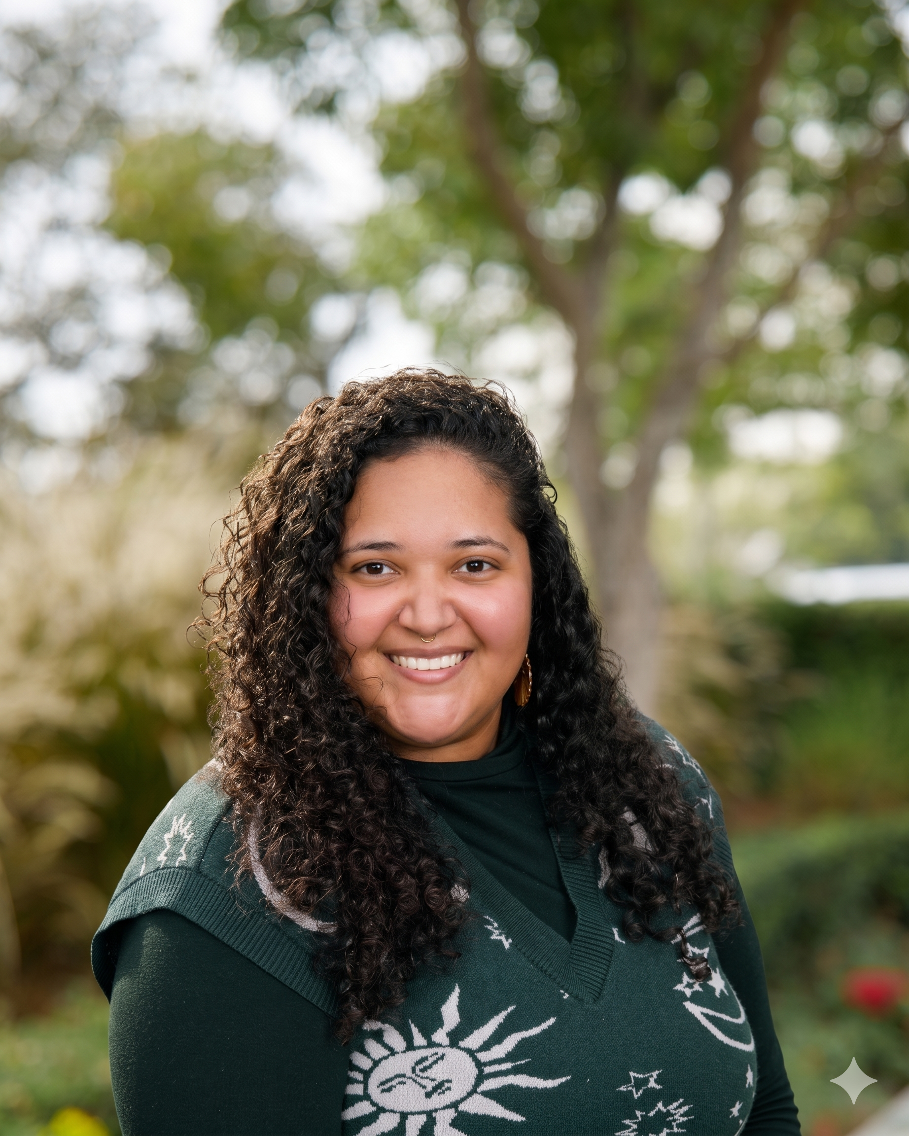Close-up of a woman with curly hair smiling outdoors, with a blurred background of green and white foliage.