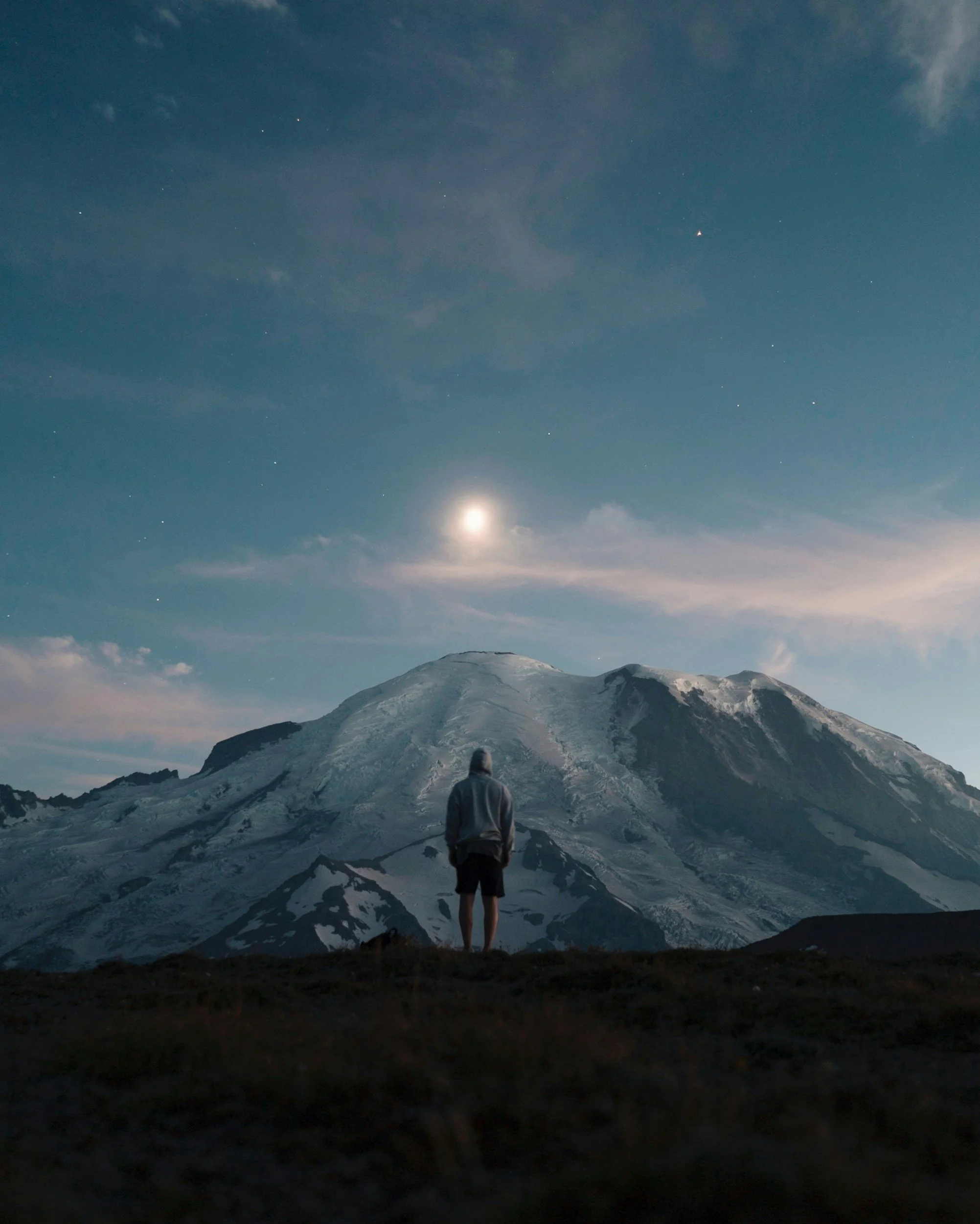 A person standing on a dark foreground with an illuminated snow-capped mountain and a partly cloudy night sky with stars and the moon.