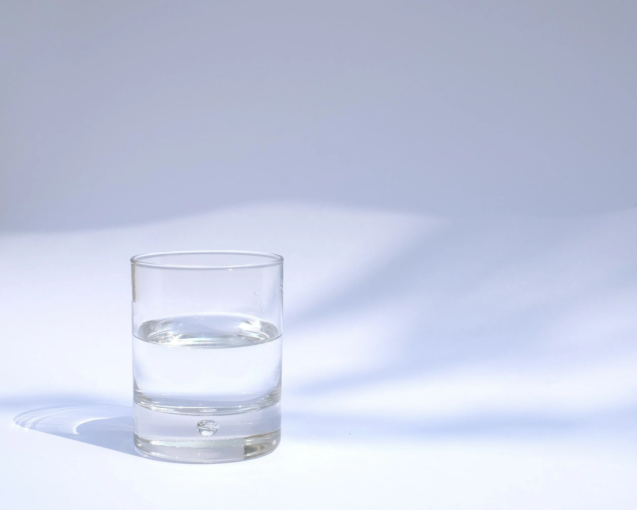 A glass of water on a white surface with a plain, light-colored background.