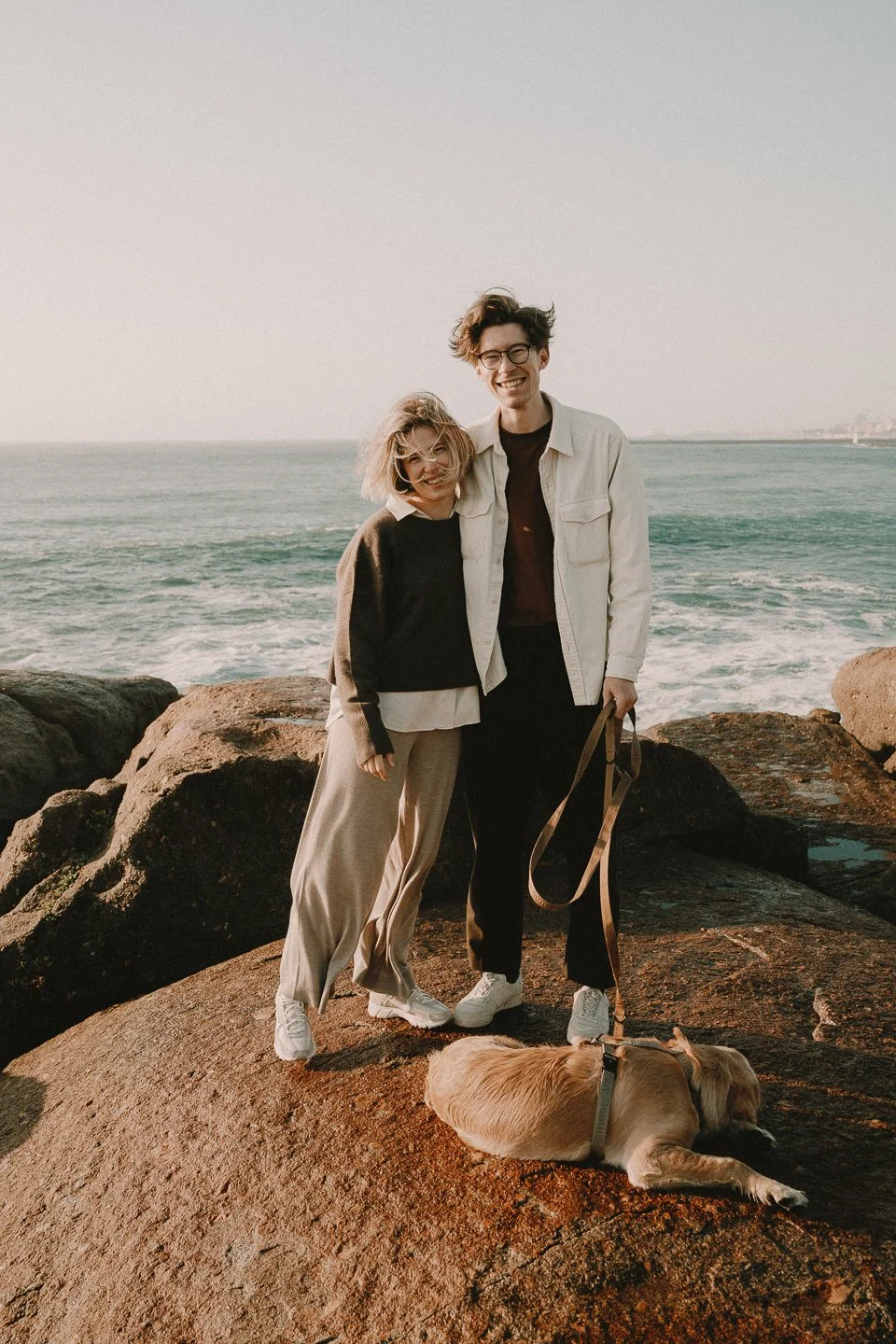 Two smiling women and a dog standing on rocks near the ocean at sunset.