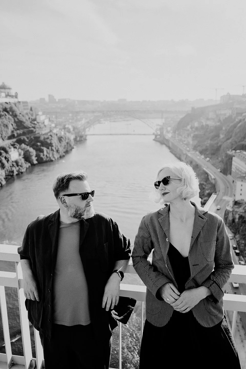 A black and white photo of a man and a woman standing on a balcony overlooking a river, with a bridge and cityscape in the background. Both are wearing sunglasses, and the woman has blonde hair styled in vintage waves.
