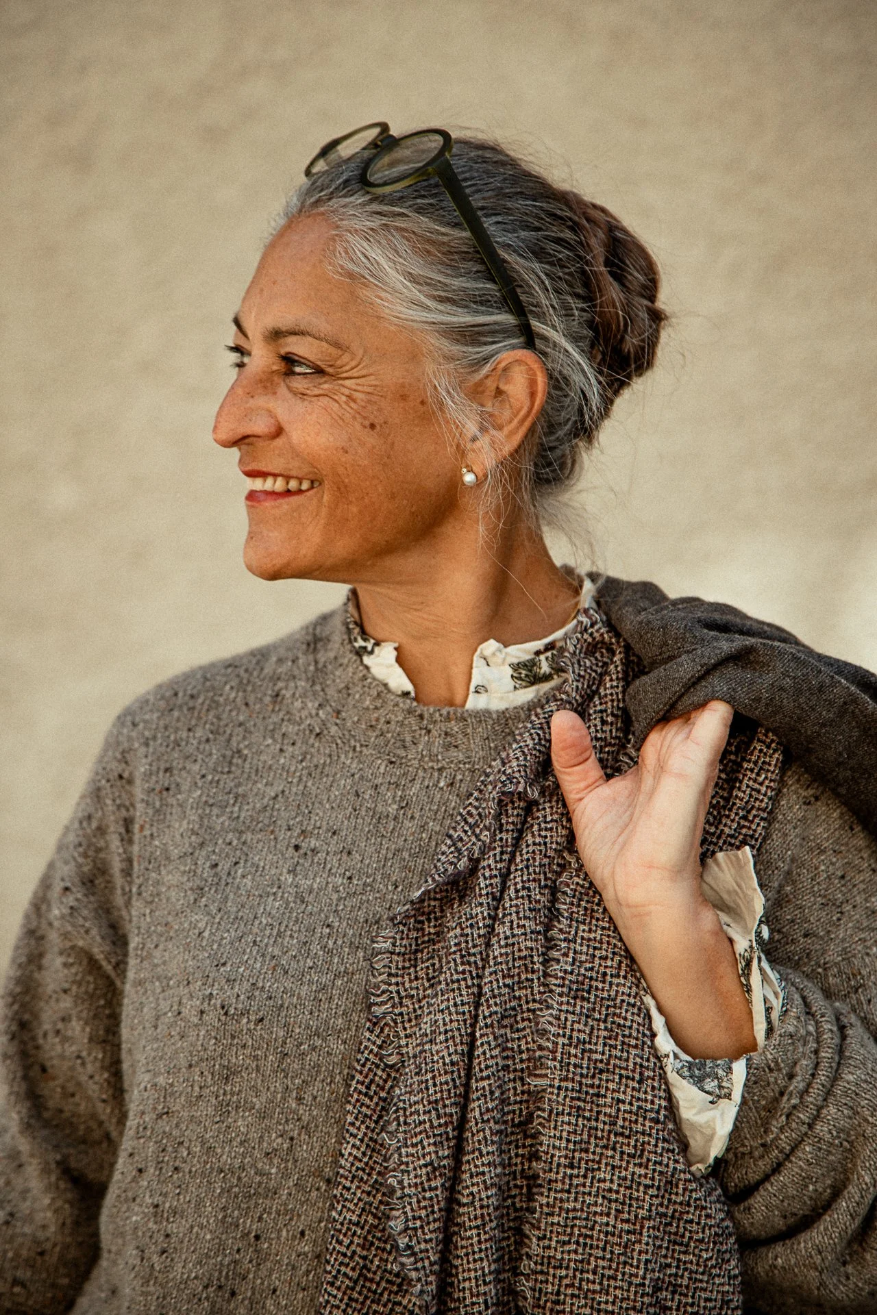 Close-up of an older woman with gray hair, glasses on her head, wearing earrings, a white blouse with floral pattern, and a sweater. She is smiling and looking to the side, holding her sweater over her shoulder.