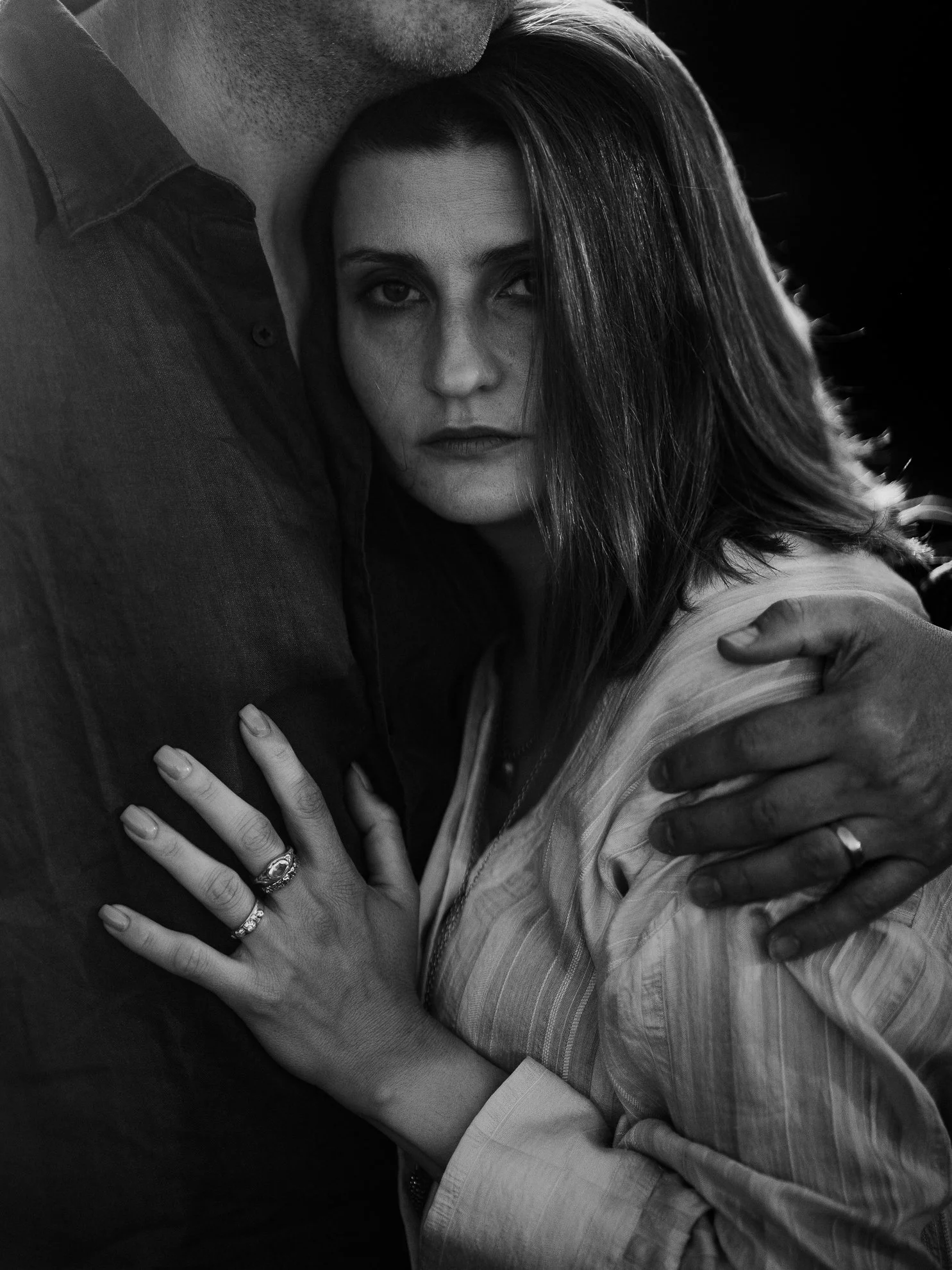 A black and white photo of a man and woman hugging. The woman's face is visible with intense eyes and a serious expression. The man is mostly out of frame, with only part of his face and shoulder visible. The woman has rings on her fingers and is wea