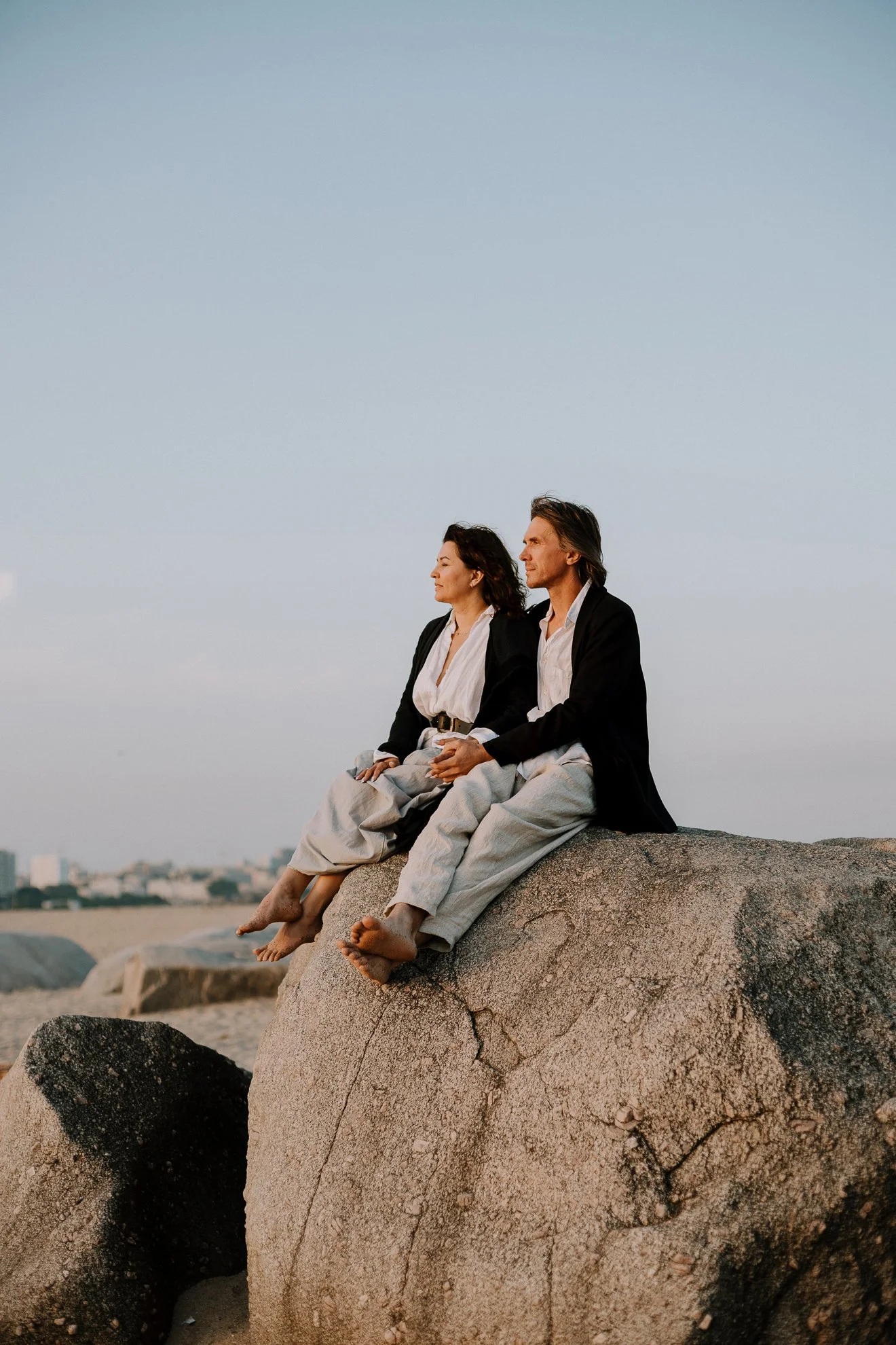 A man and woman sitting on a large rock at the beach during sunset, with their eyes closed, facing the horizon. The man has gray hair and the woman has dark hair. Both are dressed in casual, light-colored clothing.