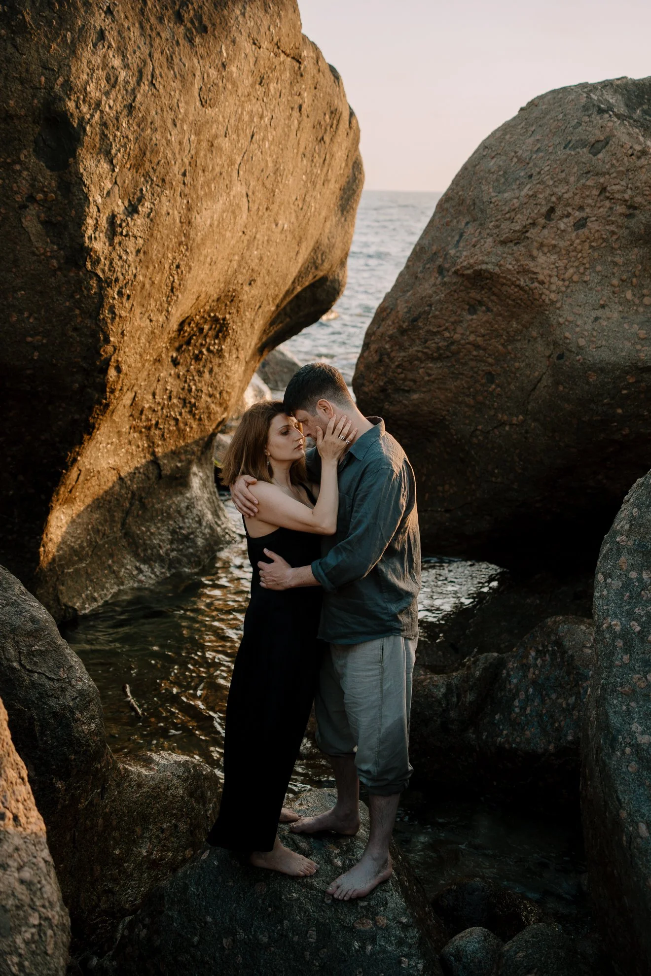 A couple stands close together with their foreheads touching, surrounded by large rocks near the water at sunset. Porto, Portugal.