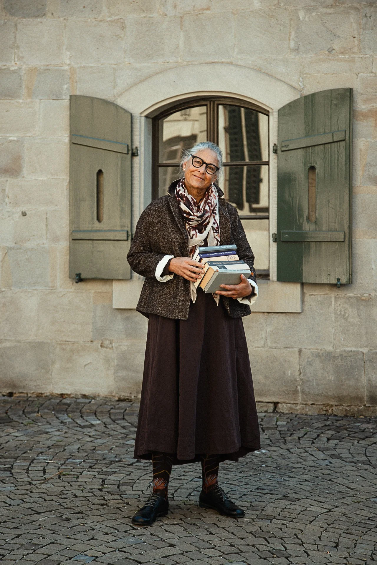 A woman standing outdoors on a cobblestone street, holding several books. She has gray hair, glasses, and is dressed in a long skirt, a blazer, and a scarf, with a neutral expression. Behind her is a stone wall with a small window with green shutters