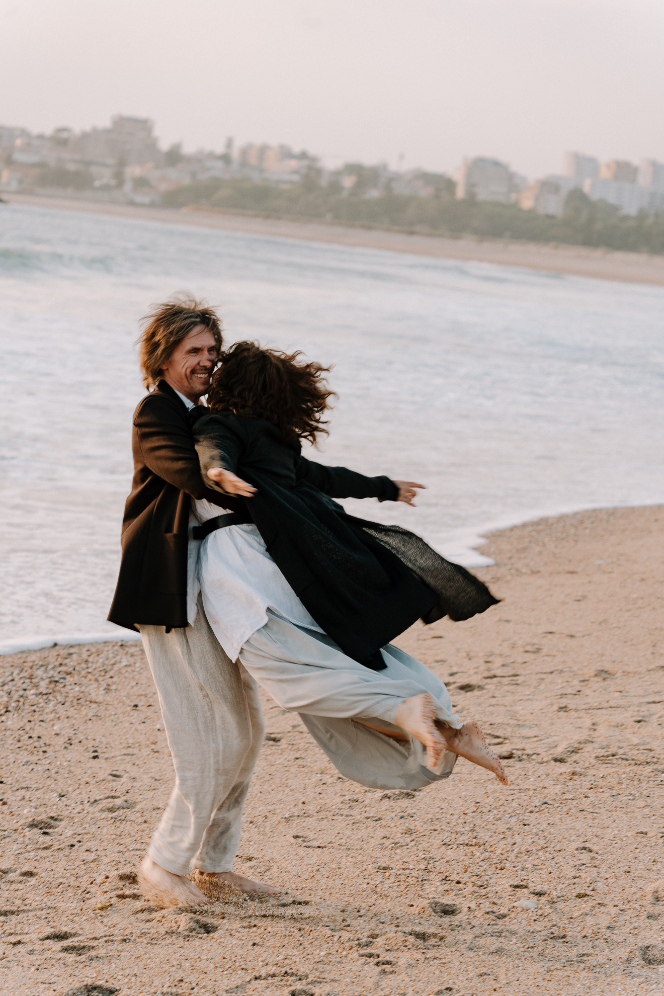 A man is holding a woman in a black coat over his shoulder on a sandy beach, with the woman laughing and kicking her legs, near the water's edge, at sunset.
