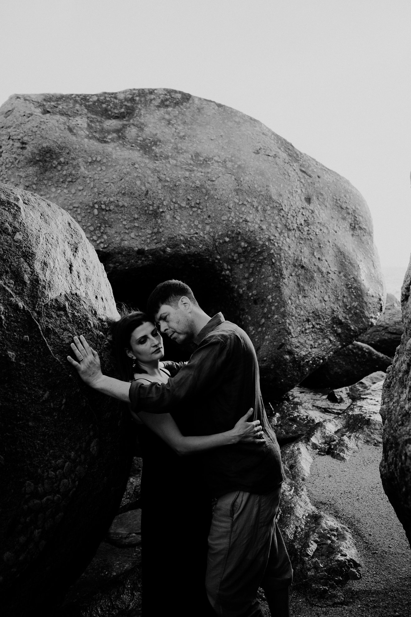 A black and white photograph of a man and a woman embracing each other among large rocks near a stream. The woman leans against a rock with her eyes closed, and the man gently holds her waist, looking down at her.