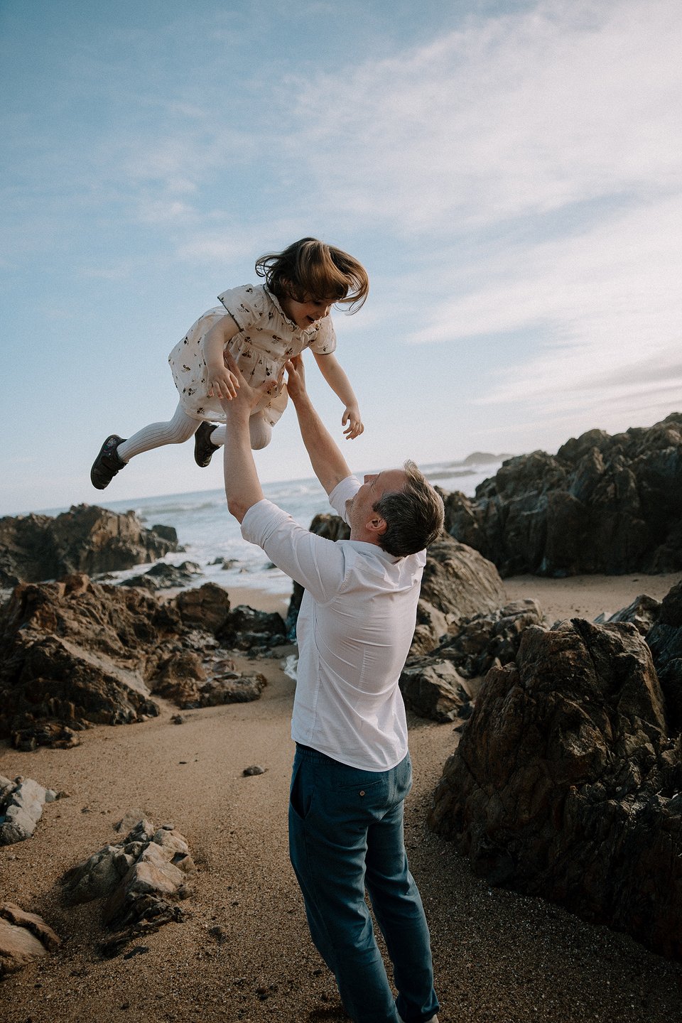 A man is lifting a young girl in the air on a rocky beach with waves and a cloudy sky in the background.