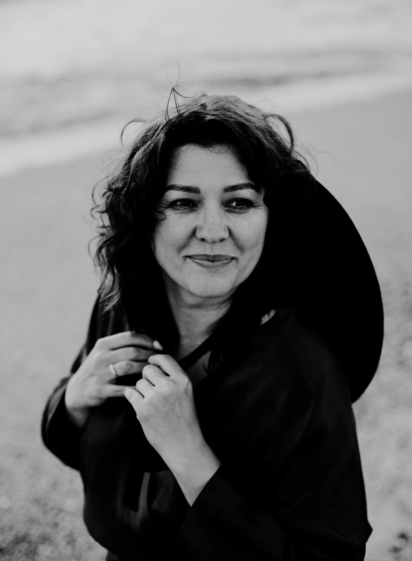 A black-and-white photo of a woman with wavy, shoulder-length hair, smiling softly, holding her hands together near her chest. She is outdoors, with a blurred background suggesting a beach or open area.