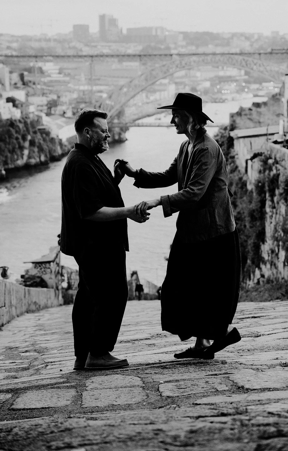 A black and white photo of a couple dancing on a cobblestone path by a river, with a cityscape and a bridge in the background.