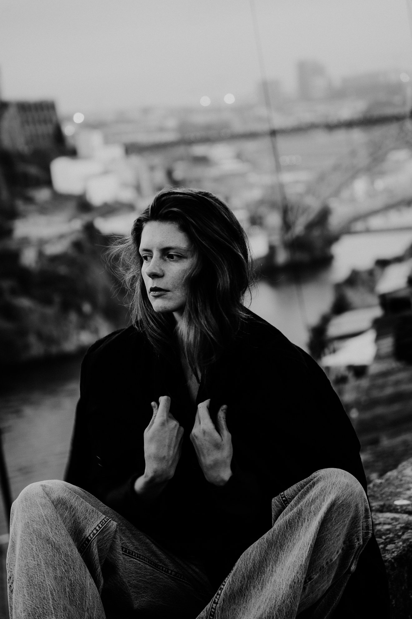 A black and white photograph of a woman sitting outdoors with a cityscape in the background. She has long hair, wears a dark top and jeans, and is looking away from the camera with her hands touching her chest.