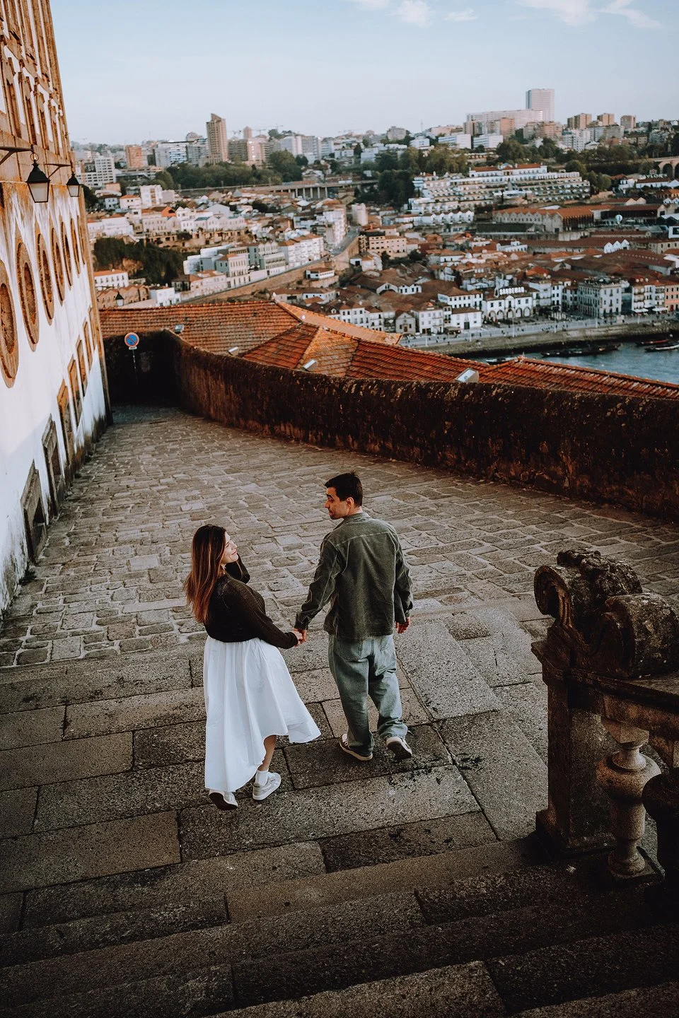 A couple holding hands and walking down stone steps with a cityscape view in the background, featuring many buildings, rooftops, and a river.