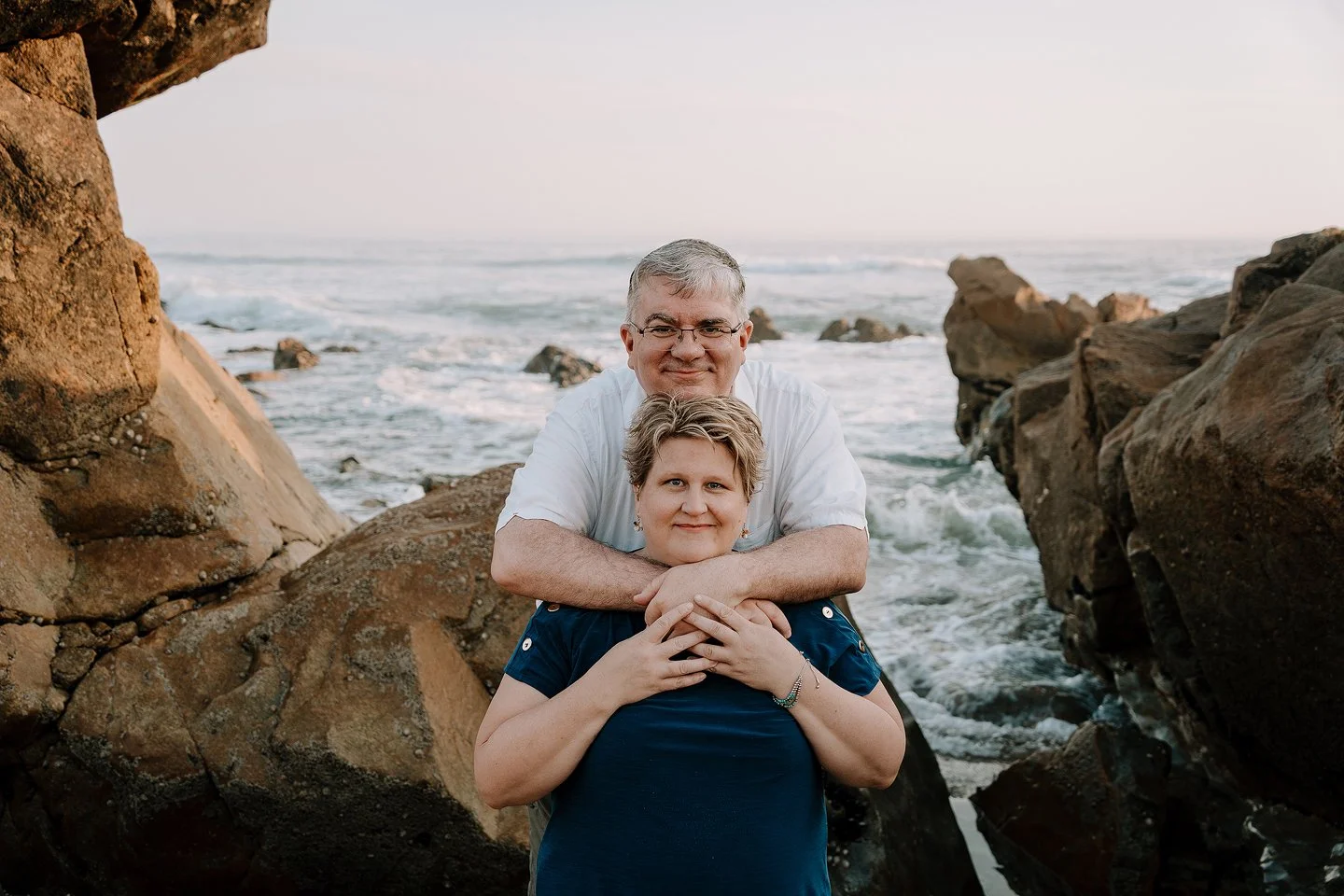 A smiling middle-aged couple with short hair, standing amidst large rocks by the ocean at sunset, embracing each other and looking at the camera.