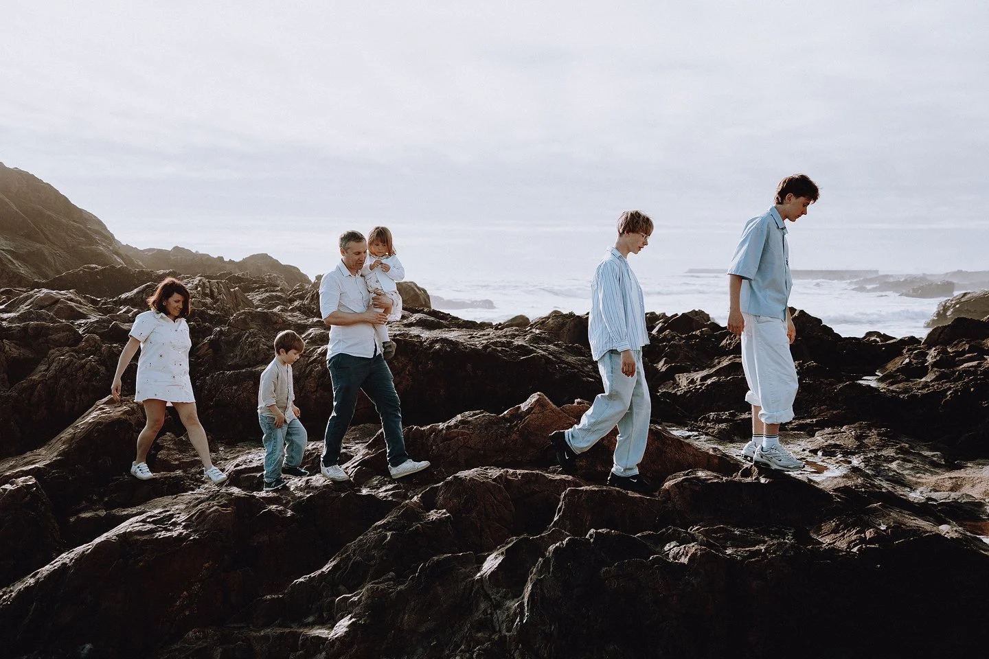 A family of six walking on rocks by the ocean, with a cloudy sky and waves in the background.