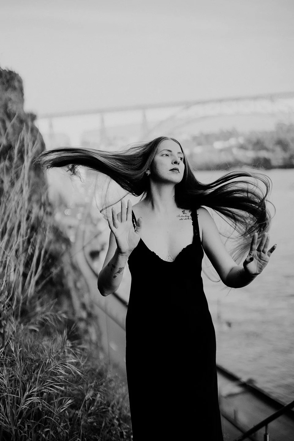 A black and white photo of a young woman with long hair, wearing a sleeveless black dress, standing by water with a bridge in the background. She has tattoos on her arms and is holding her hands up with open palms.