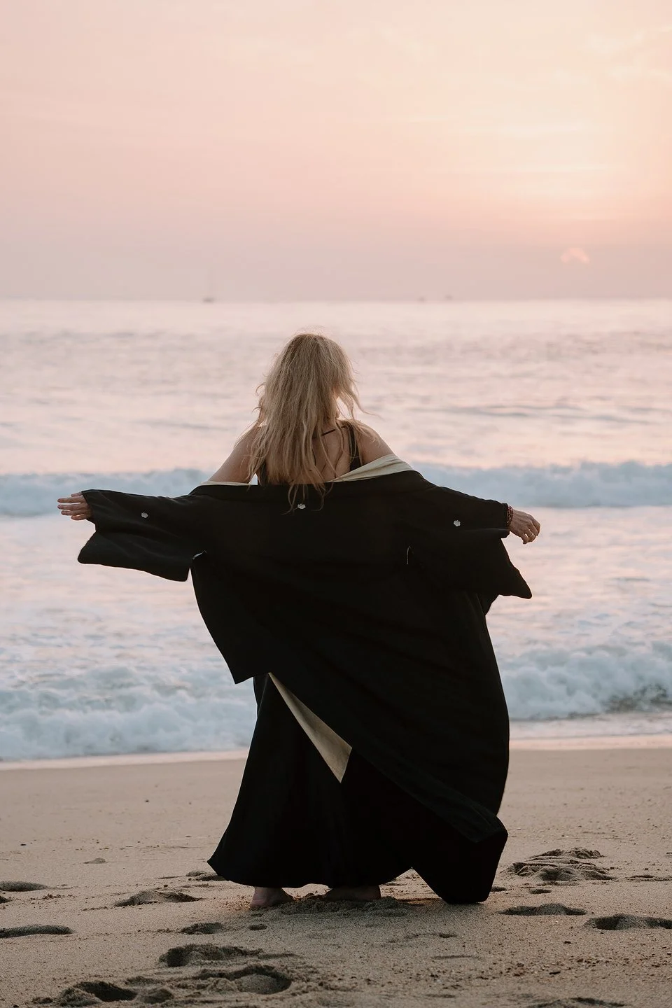 A woman with blonde hair standing on the beach at sunset, holding a black coat open while facing the ocean.