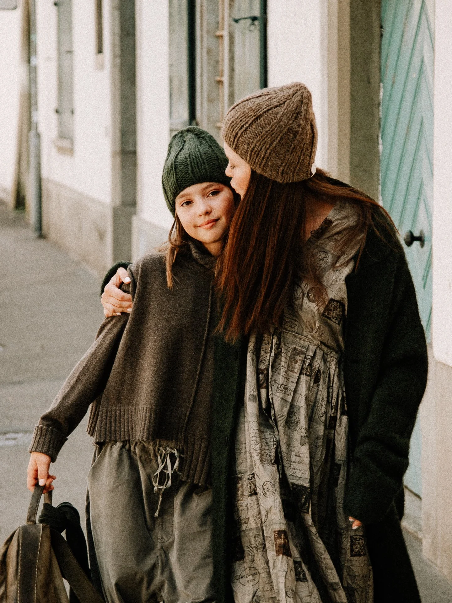 A woman and a young girl hugging on a city street, both wearing knit beanies and casual clothing, with buildings in the background.