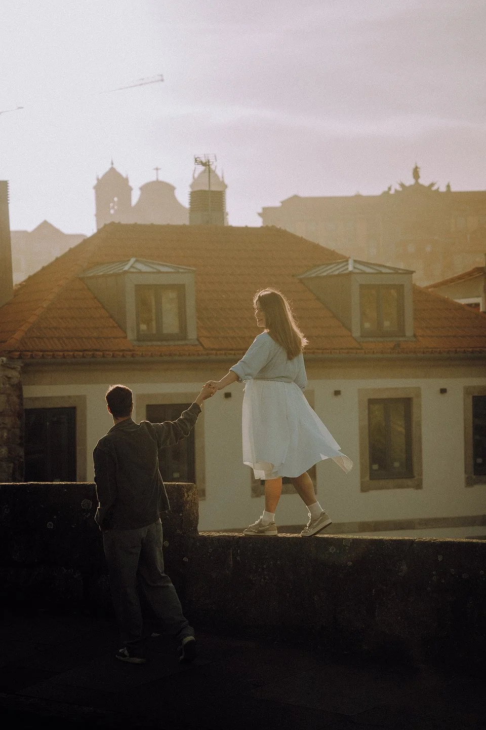 A young woman in a white dress holding hands with a young man in a dark jacket, standing on a rooftop during sunset with city buildings in the background.