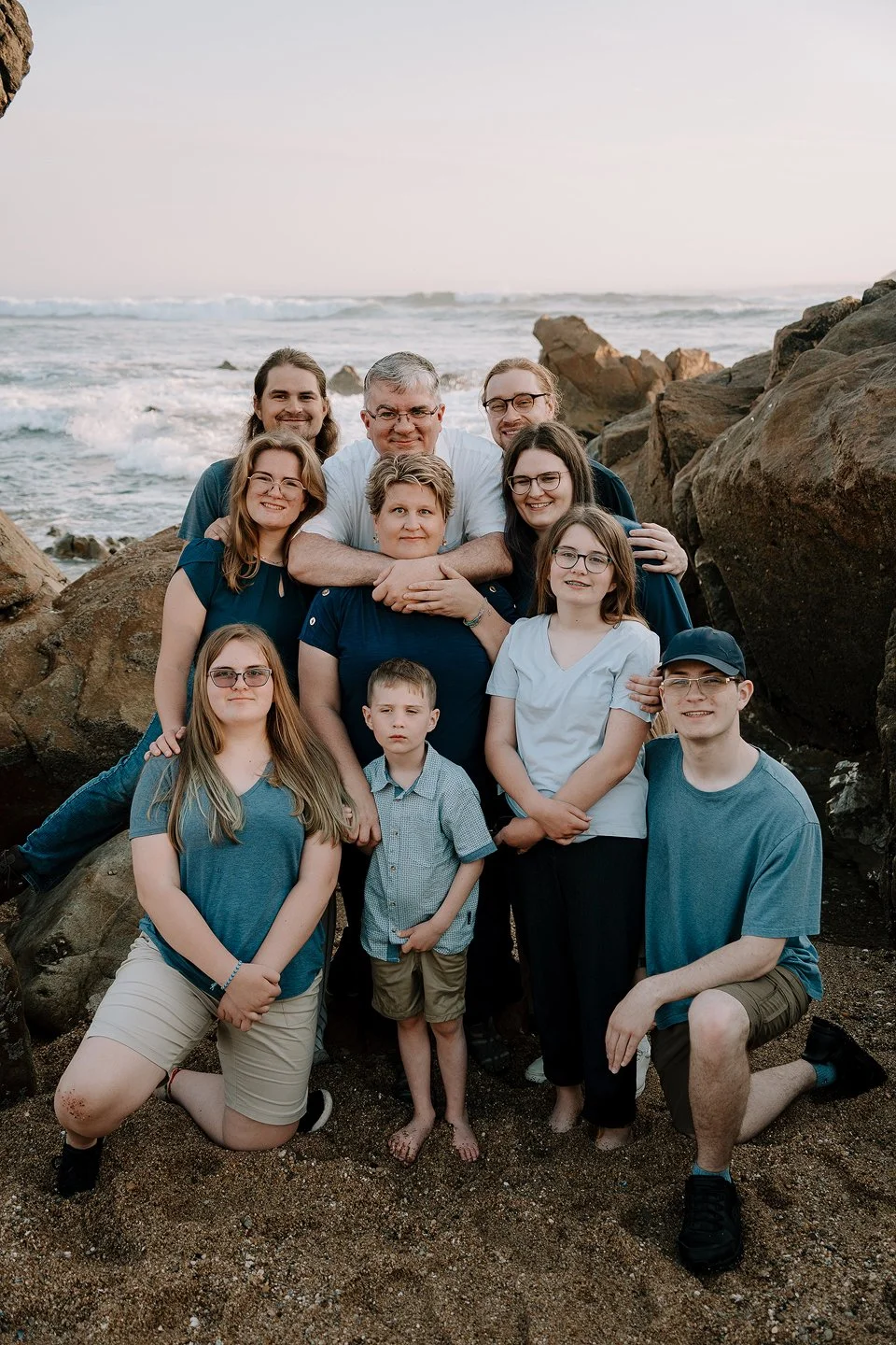 A large family group of ten people is posing together on a beach with rocks and ocean waves in the background. The group includes adults and children, all smiling and dressed casually for the beach.