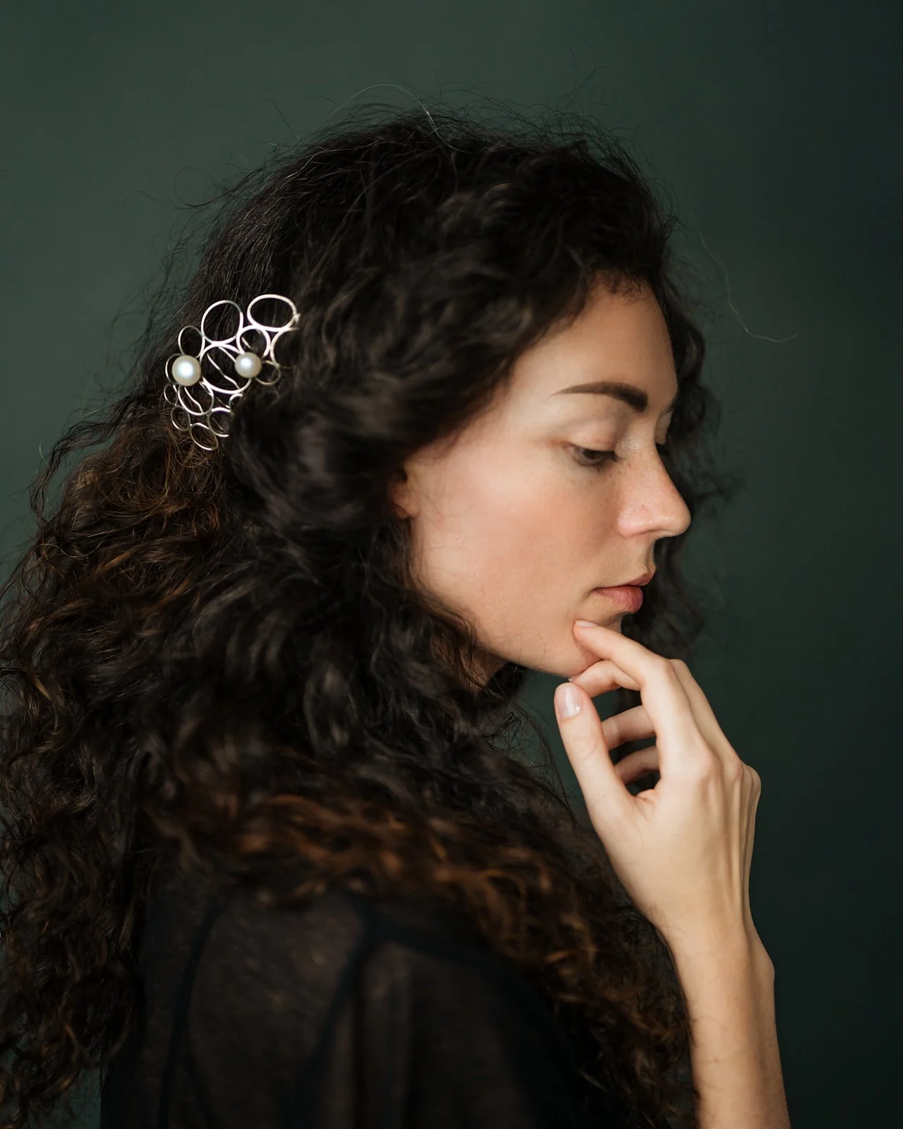 A woman with dark curly hair wearing a decorative hair accessory with pearls, touching her chin, against a dark green background.
