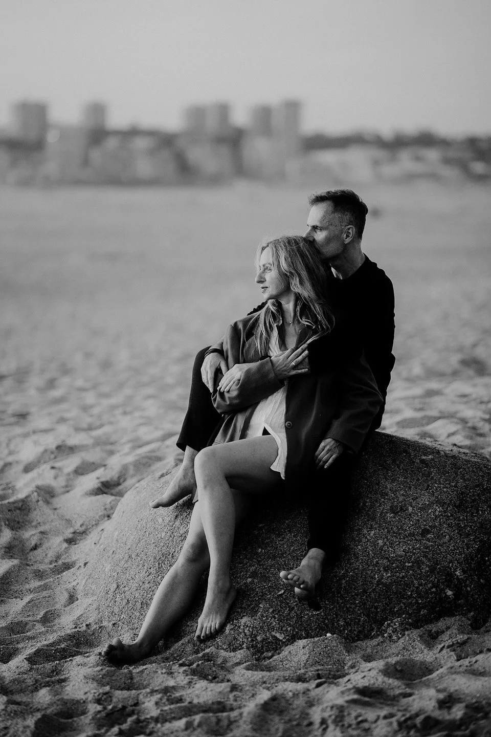 A black-and-white photo of a couple sitting on a large rock on a beach. The woman has long, wavy hair and is looking to the side, while the man is behind her, kissing her on the forehead. The background shows blurred buildings in the distance.