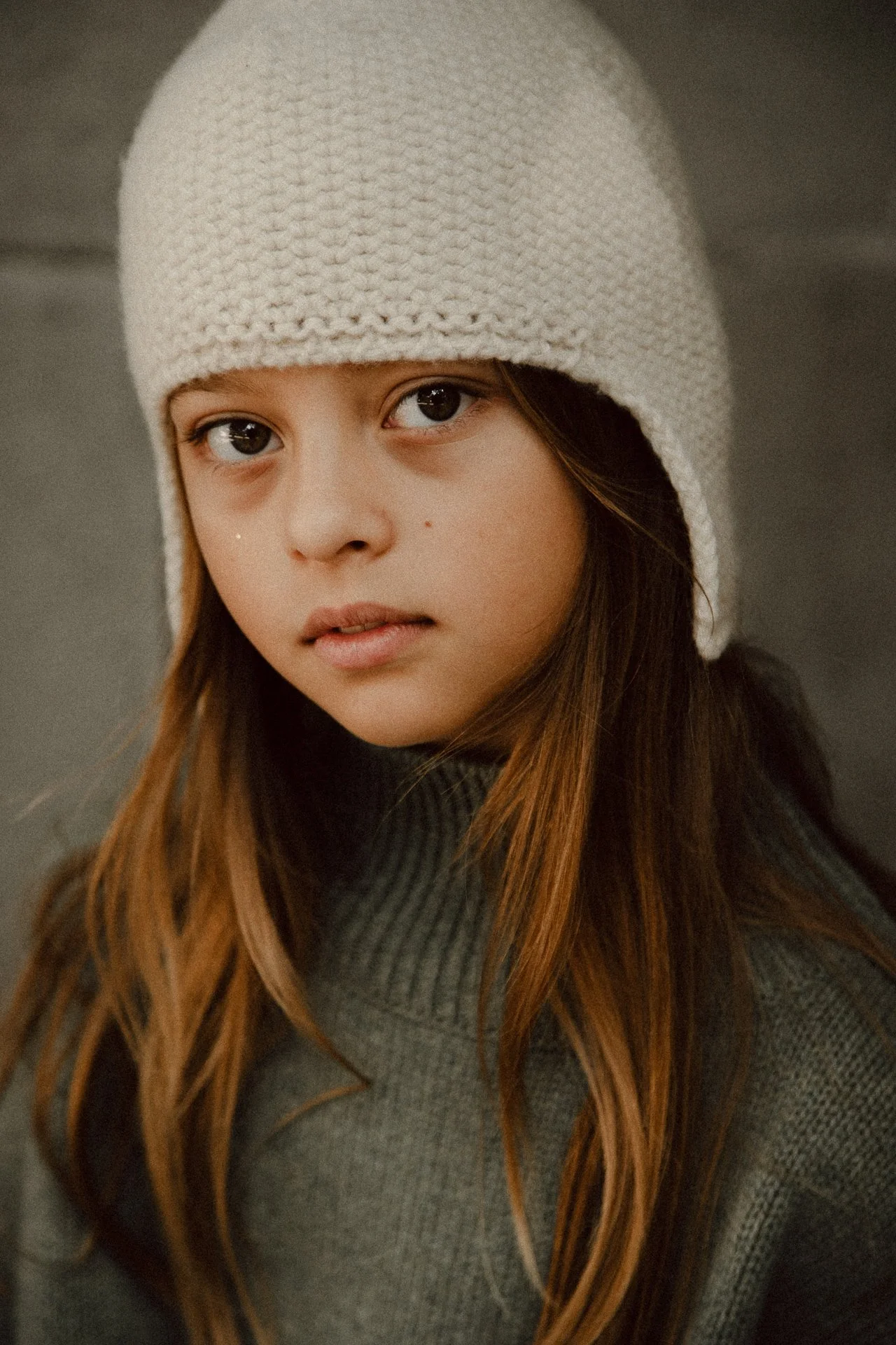 Close-up of a young woman with long brown hair, wearing a white knit hat and a gray sweater, looking at the camera.