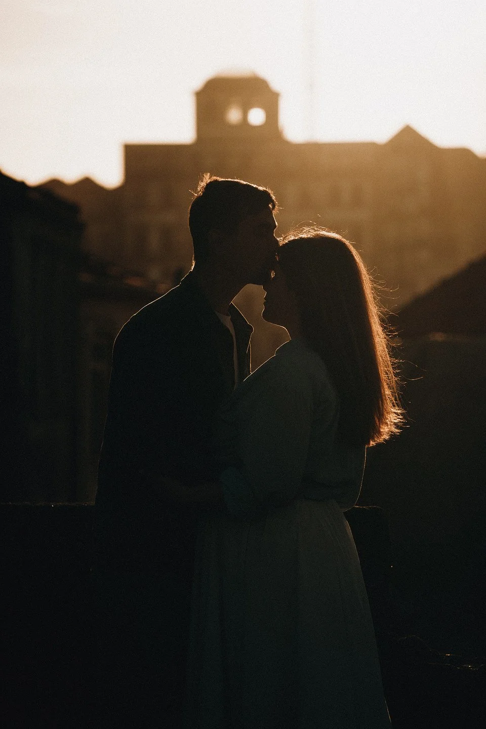 A silhouette of a couple kissing in front of a sunset with a building in the background.