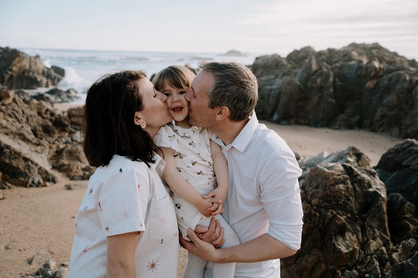 A family of three at the beach, with a woman and man kissing their young daughter on the cheeks. The ocean and rocks are in the background.