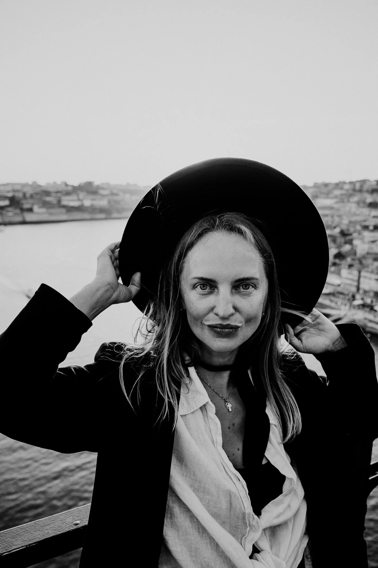 A woman with long hair wearing a fedora hat, white shirt, and dark blazer, sitting outdoors near water with a cityscape in the background, holding her hat with one hand, smiling slightly.