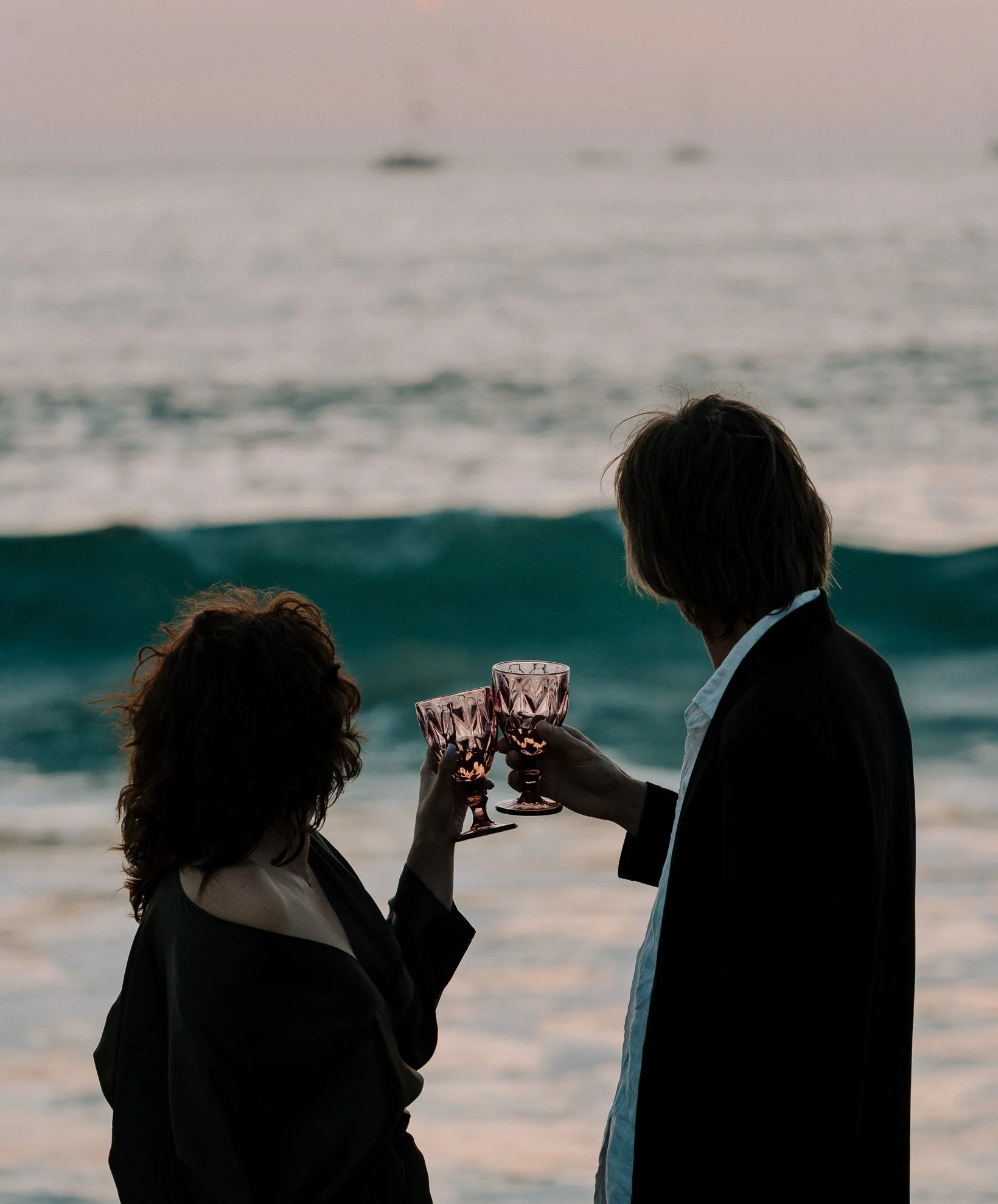 Two people in formal attire at the beach, toasting with pink wine glasses during sunset.