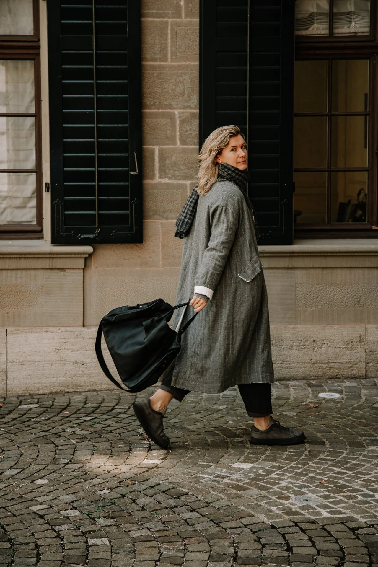 A woman walking on a cobblestone street, pulling a black bag, wearing a long gray coat, black pants, and brown shoes, with a black and gray scarf, in front of a brick building with black shutters and windows.