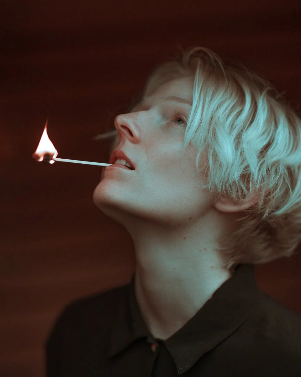 A woman with short, blonde, wavy hair holding a lit matchstick in her mouth, with a dark background.