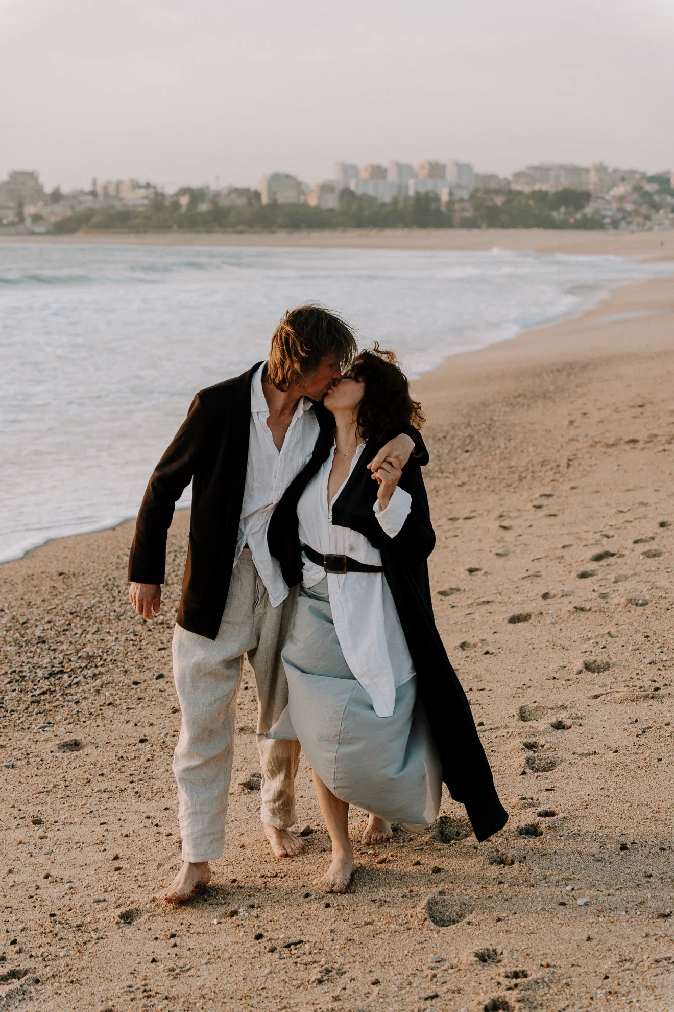 A man and woman sharing a kiss on a sandy beach during sunset, with city buildings in the background.