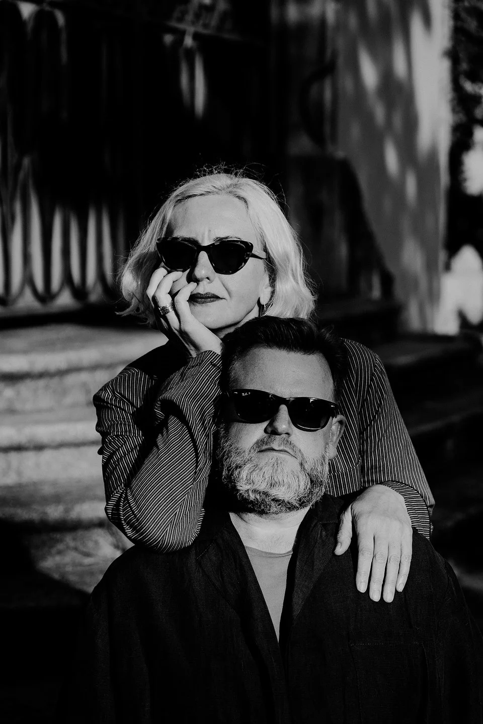 Black and white photo of an older woman with blonde hair and an older man with a beard, both wearing sunglasses. The woman is leaning over the man with her hand on his shoulder, outdoors in sunlight.