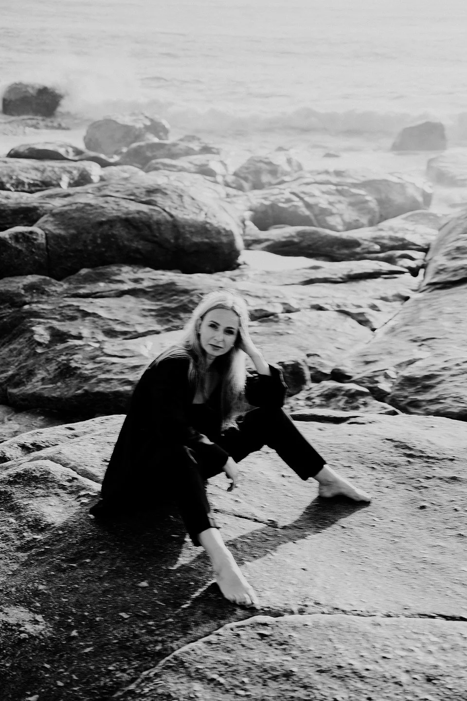 A woman sitting on a rocky shoreline near the ocean, in black and white.