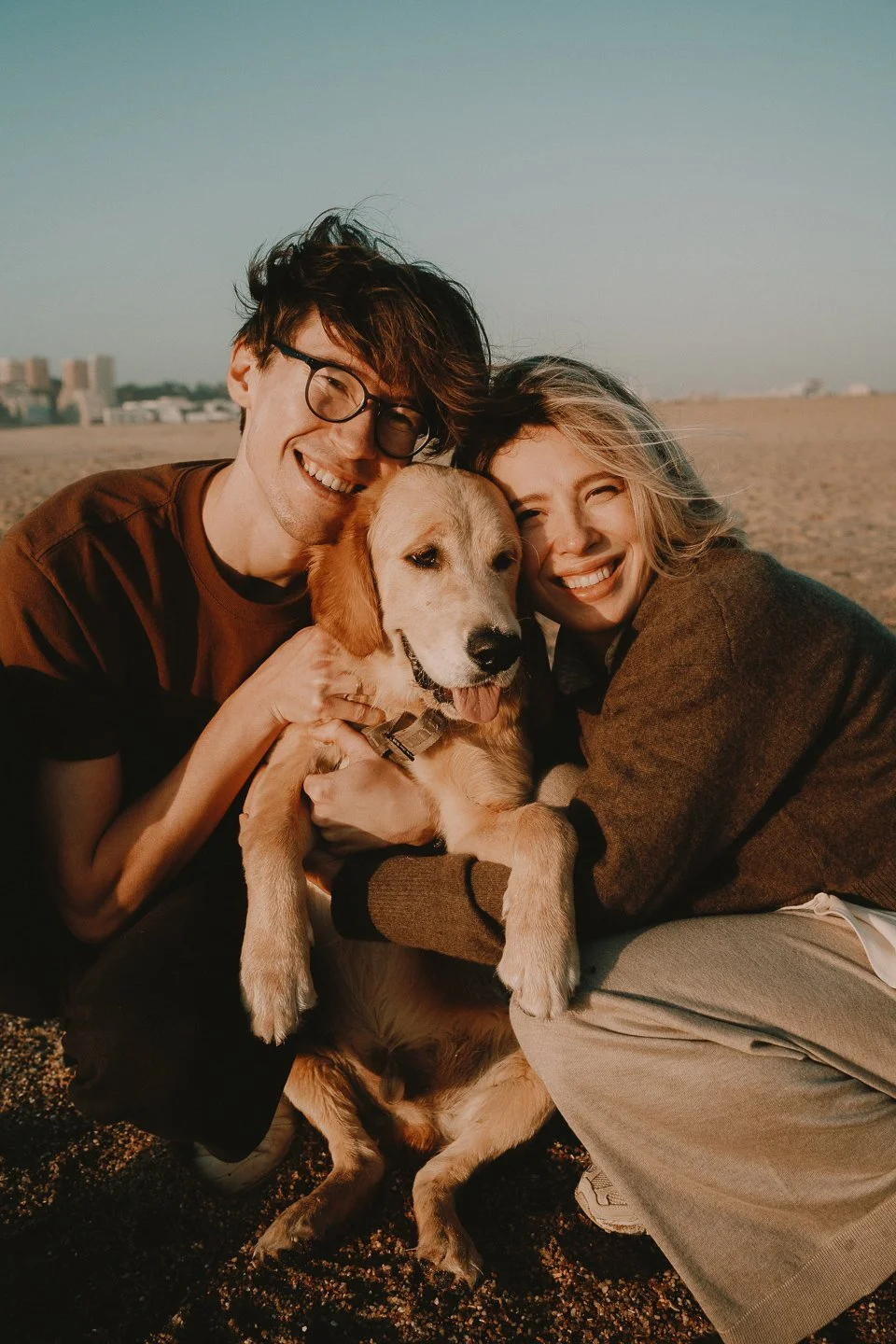A happy couple hugging a Golden Retriever puppy at the beach during sunset.